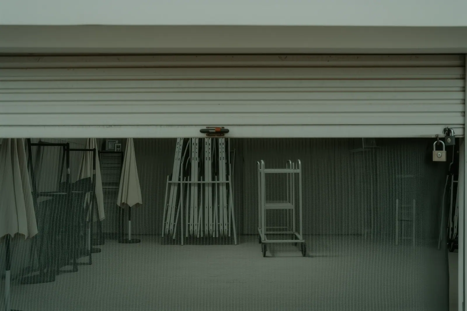 Stacked sun loungers, folded parasols and a hand cart inside a locked beach equipment store, the half-raised mesh shutter and a padlock hasp in front.