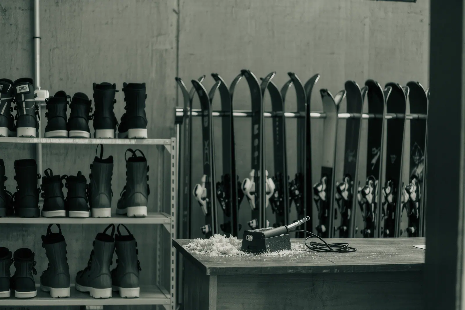 Ski and snowboard racks in a coastal rental shop with boots lined up on a steel shelf and a workbench holding a waxing iron and wax scrapings.