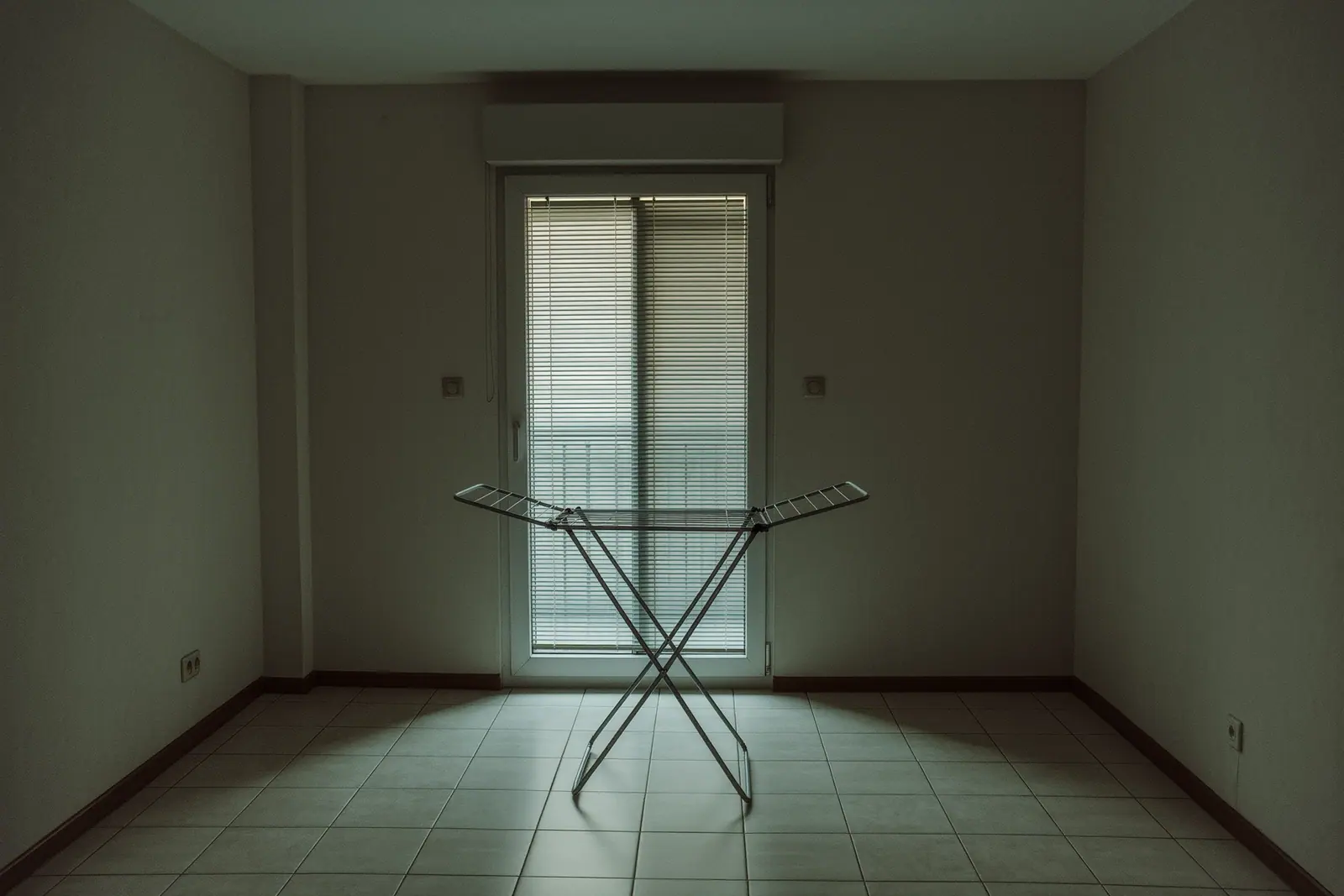 An empty long-let apartment with tiled floors and a shuttered balcony door, a bare folding drying rack standing open in the middle of the room.