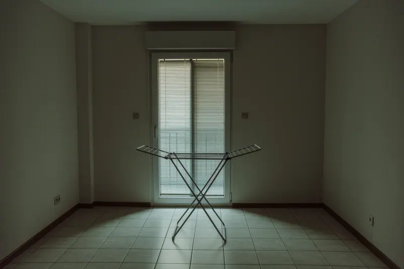 An empty long-let apartment with tiled floors, a shuttered balcony door standing ajar and a bare folding drying rack against the wall.