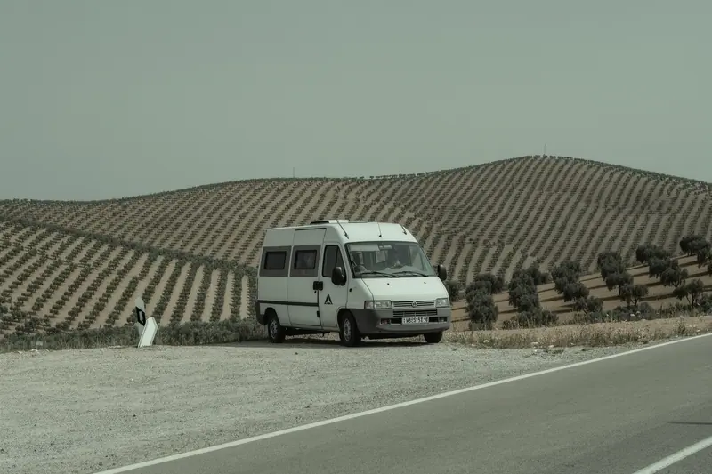 A compact campervan on a gravel pull-in beside an inland Andalusian road, olive terraces rising behind it.