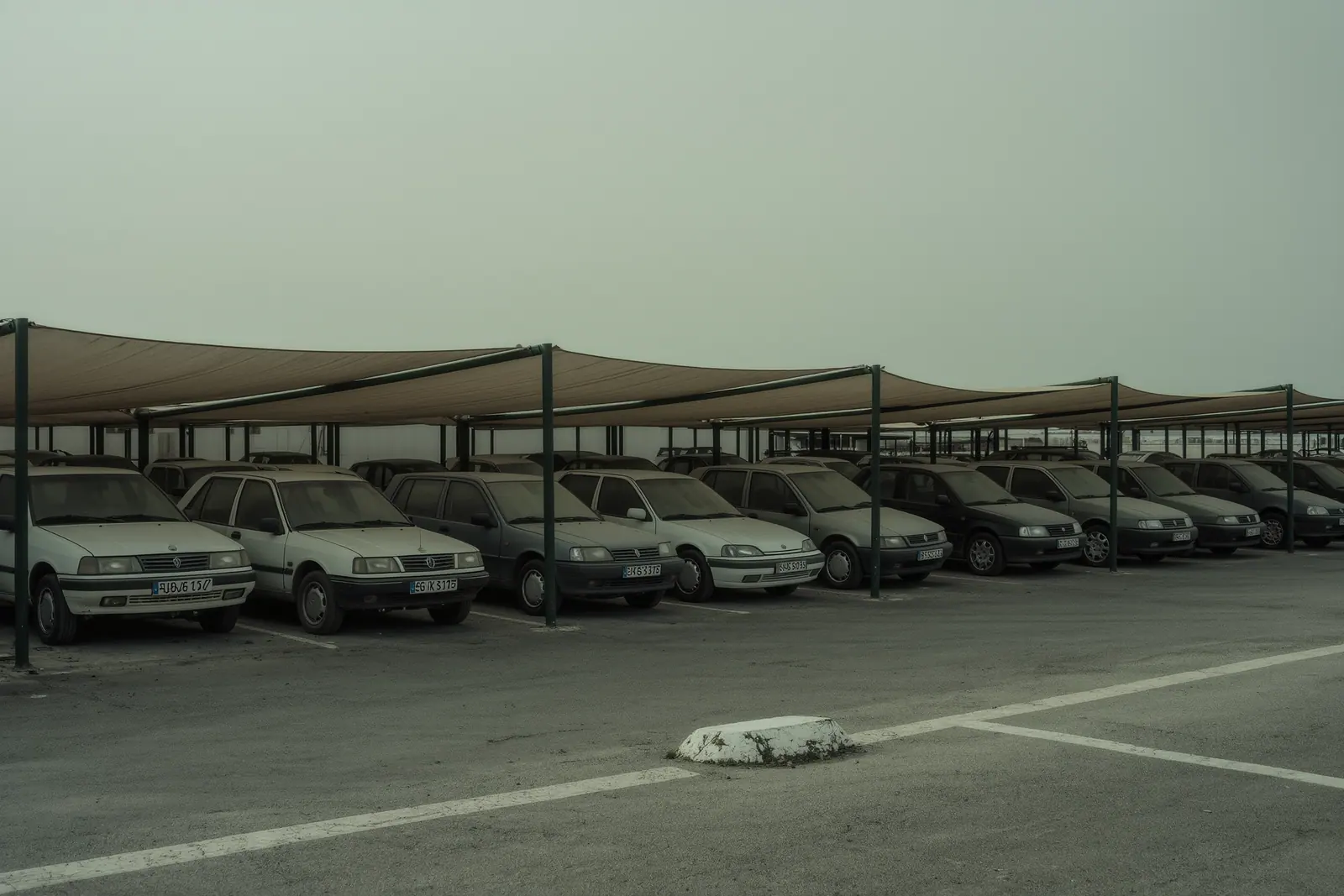 A quiet long-stay parking lot on the Costa del Sol with dusty cars standing under thin shade sails, a faded bay line and a cracked concrete wheel stop in the foreground.