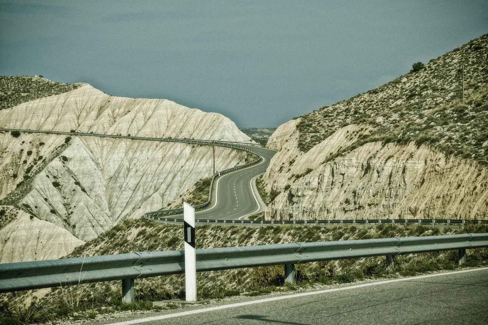 An empty mountain road switchbacking up dry limestone country towards Ronda, a galvanised crash barrier and a plain white kilometre post at the roadside.