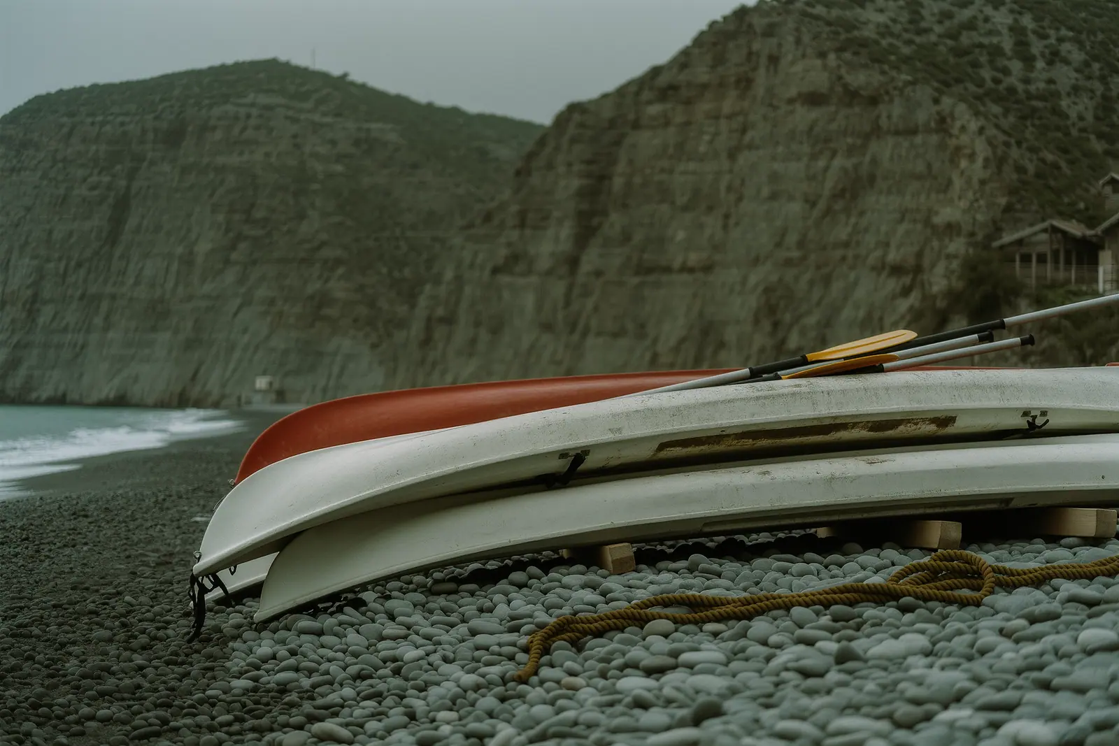 Sea kayaks and paddles stacked on a pebble beach below the cliffs near Nerja, a scuffed hull and a length of rope lying over the pebbles.