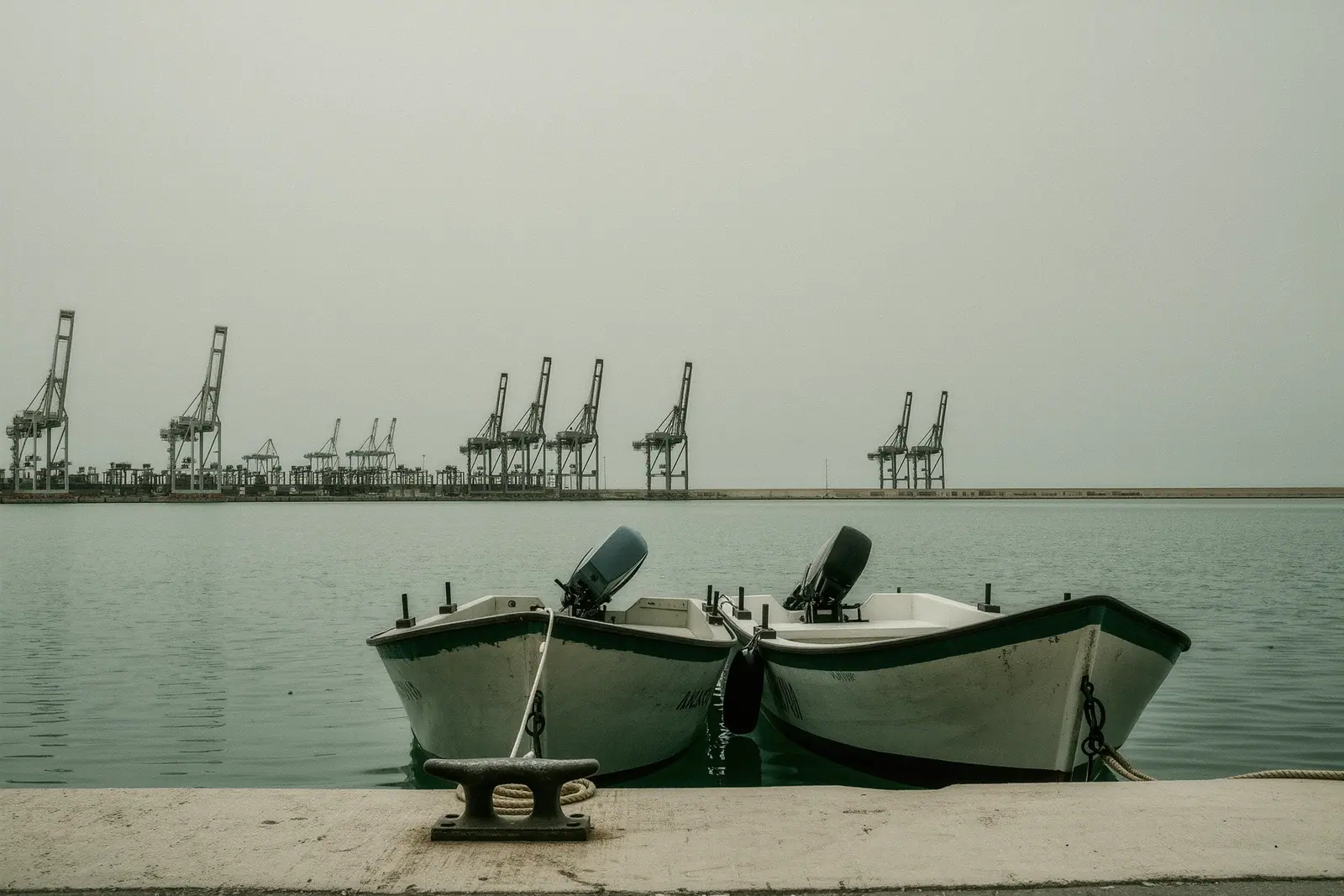 Small licence-free motorboats tied bow-to at a floating pontoon in Malaga port, a cleat and coiled painter on the decking with harbour cranes flat and grey behind.