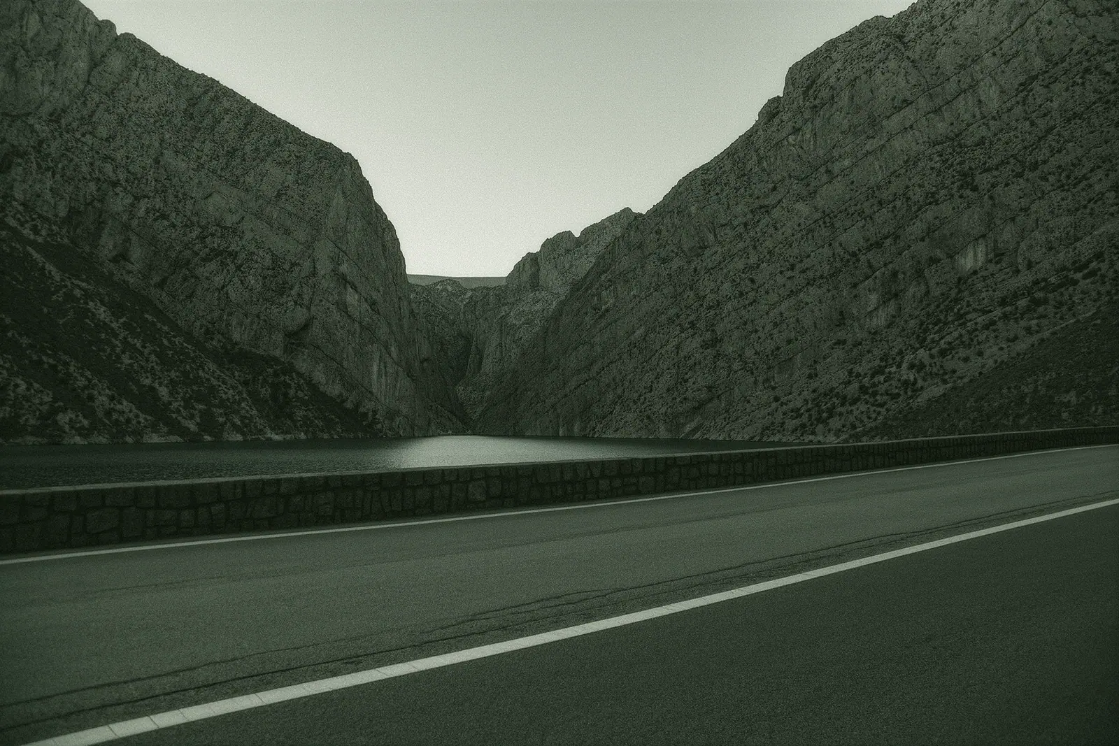 The narrow gorge approach road near El Chorro with sheer rock walls and a still reservoir below, empty tarmac and a white painted edge line running into the frame.
