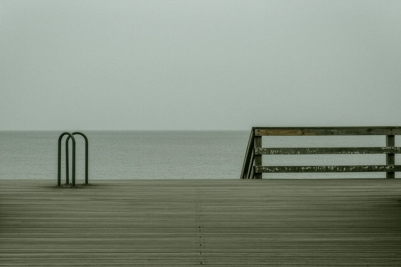 An empty boardwalk section of the coastal path with a steel bike rack and a weathered wooden handrail, timber deck boards running away from the camera and flat grey sea beyond.
